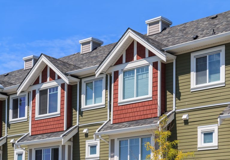 Close-up of a gray architectural shingle roof with white-trimmed gabled dormers on green and red townhomes.