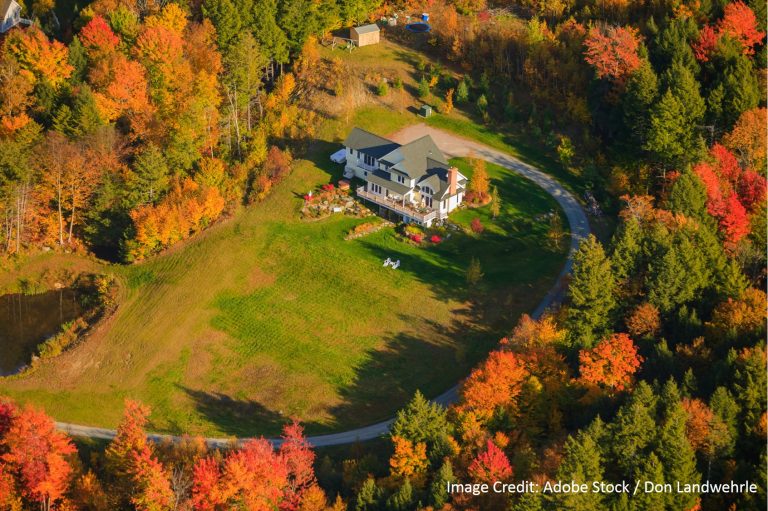 An aerial drone view shows a large white home with a dark shingle roof surrounded by vibrant autumn foliage.