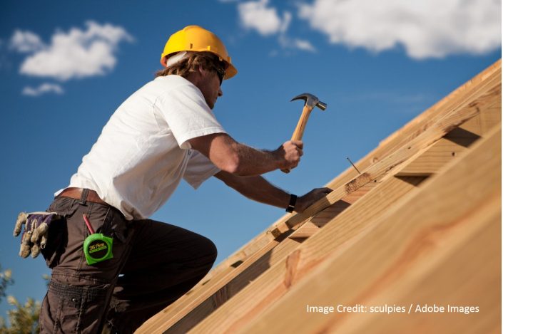 A workerin a yellow hard hat hammers nails into wooden roof decking against a bright blue sky.
