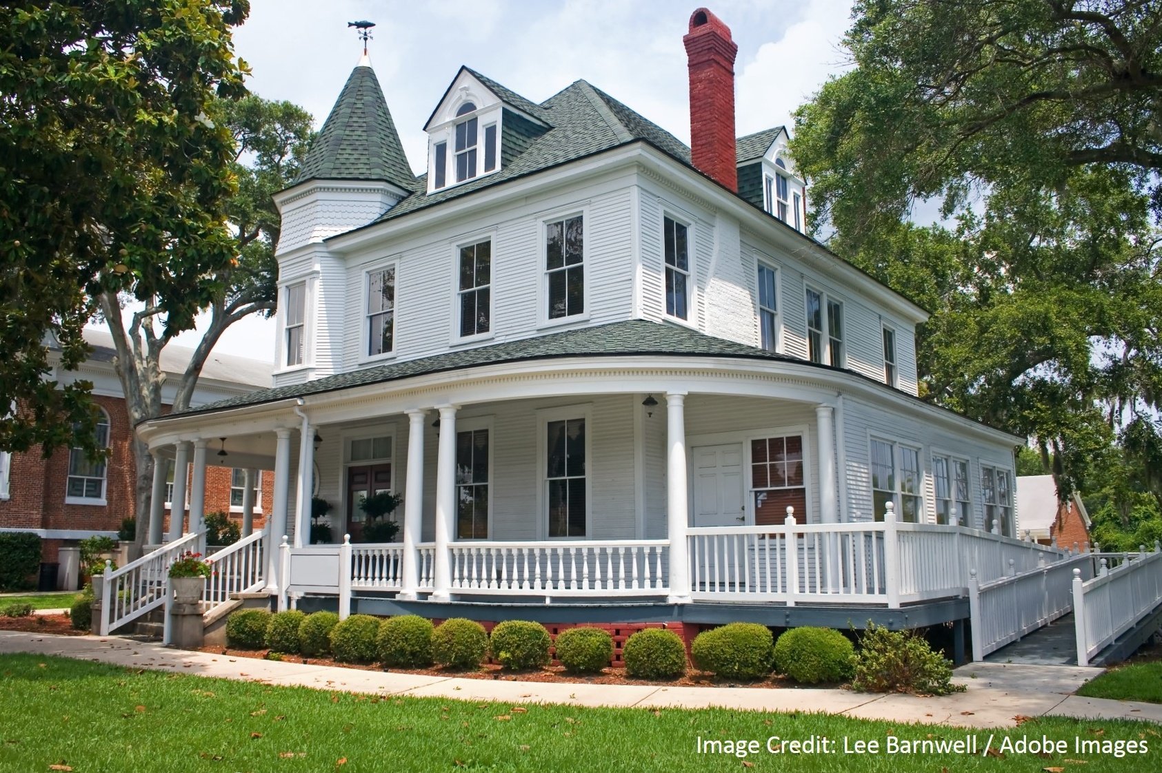 A white victorian-style home with a turret, red chimney, and wraparound porch sits behind a landscaped front yard.