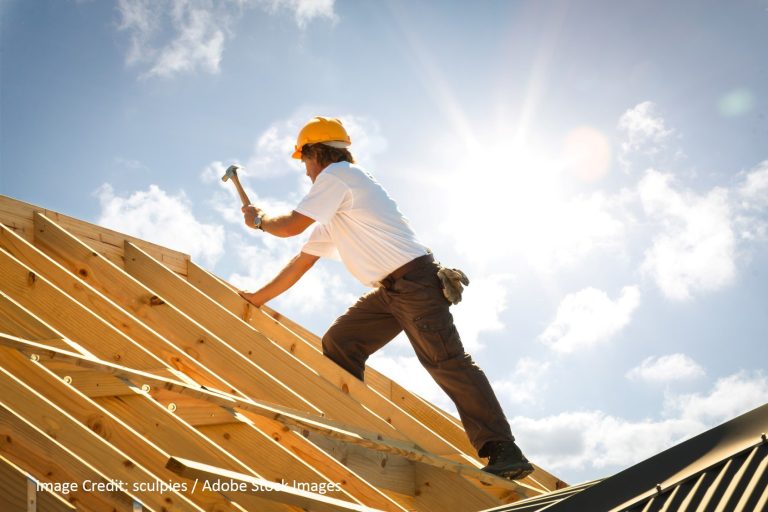 A worker in a yellow hard hat climbs exposed wooden roof trusses with a hammer against a sunlit sky.