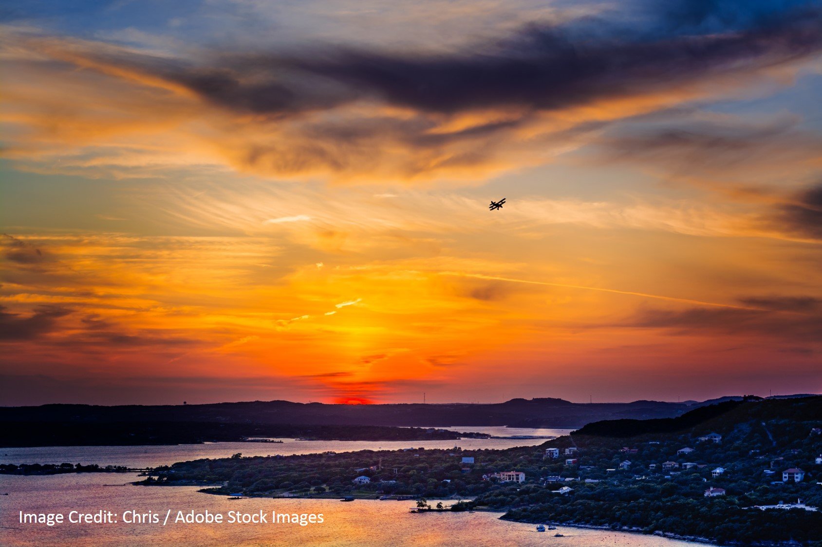 Dramatic orange and purple sunset over a lake, with wooded hills, shoreline homes, and a small plane in the sky.