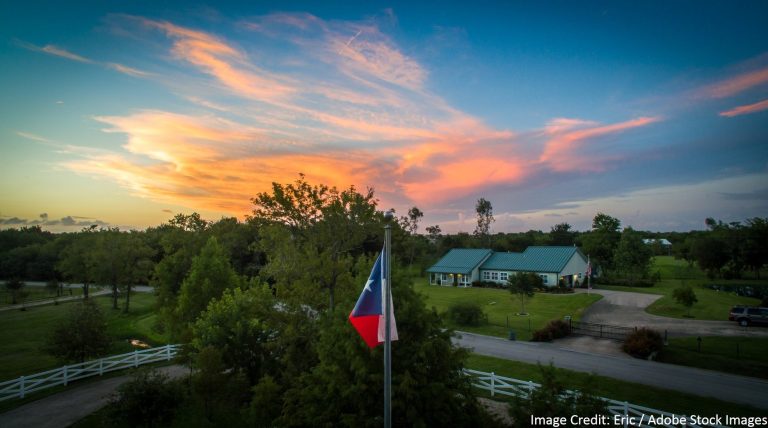 Texas flag flying on a pole with a rural home and green metal roof in the background during a colorful sunset in Bastrop.