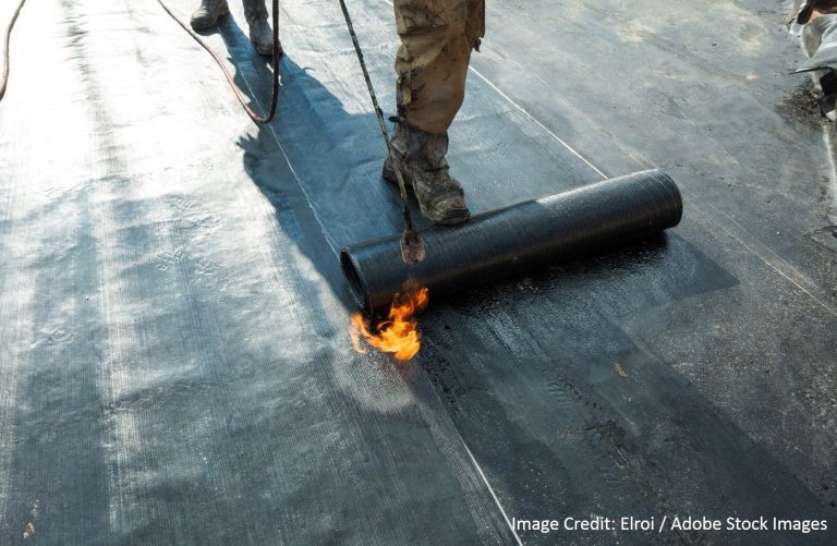 A worker uses a propane torch to heat-seal a rolled black roofing membrane onto a flat rooftop surface.