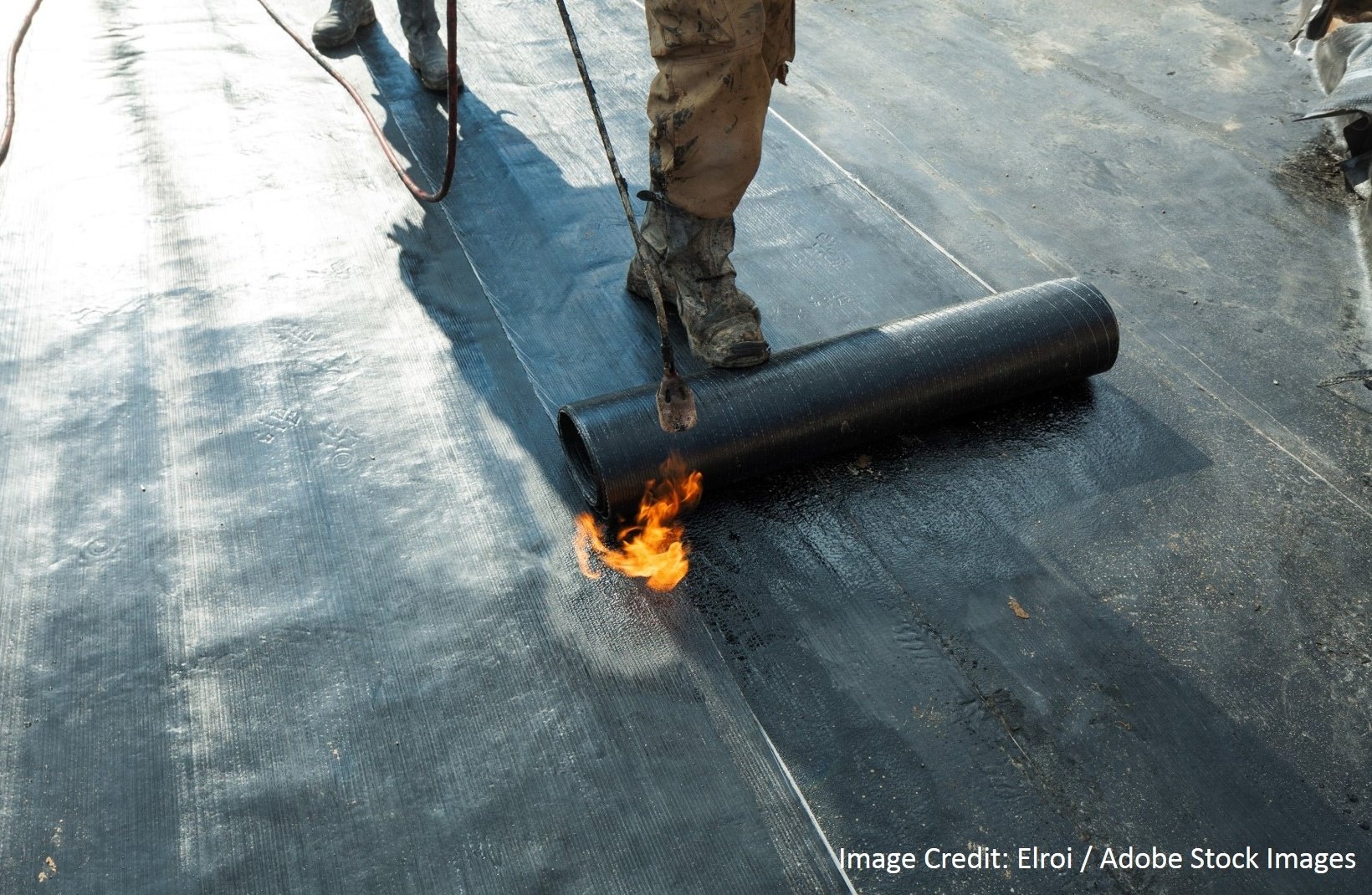 A worker uses a propane torch to heat-seal a rolled black roofing membrane onto a flat rooftop surface.