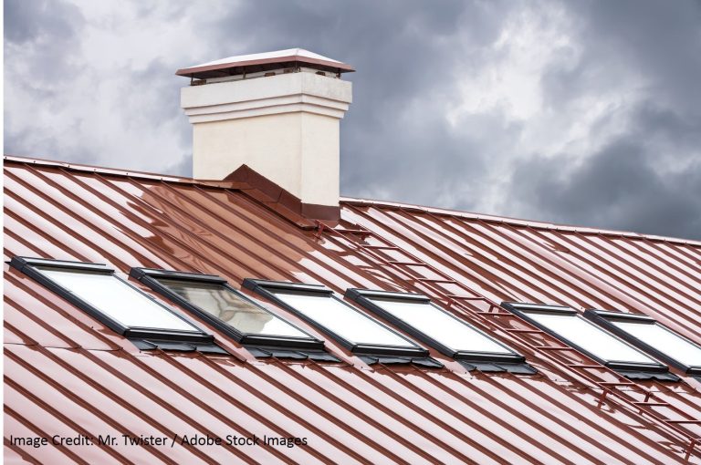 Red standing seam metal roof lined with angled skylights and a white stucco chimney against a dramatic overcast sky.