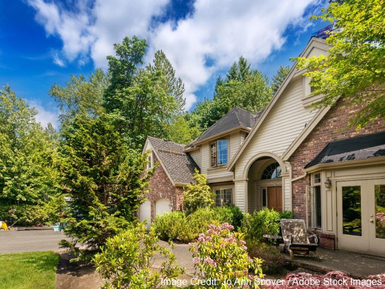 A large brick and siding home with an arched entry and dark shingle roof sits behind flowering shrubs and trees.