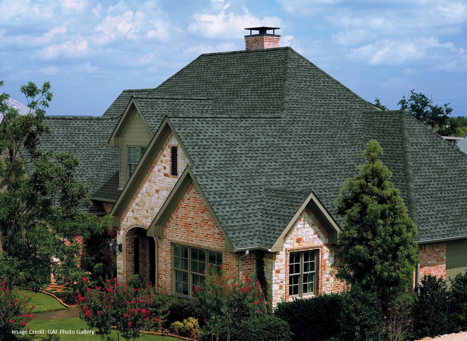 A brick home with a green dimensional shingle roof and multiple gables sits behind a landscaped flower garden.