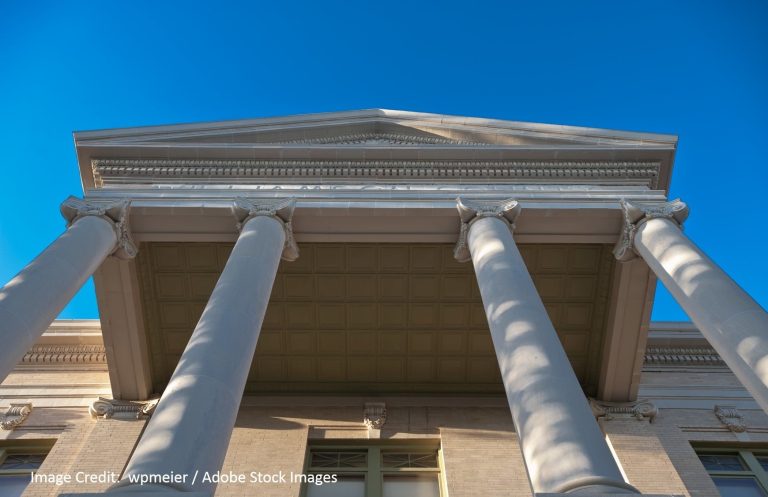 A low-angle view of tall classical columns supporting a triangular pediment against a bright blue sky.