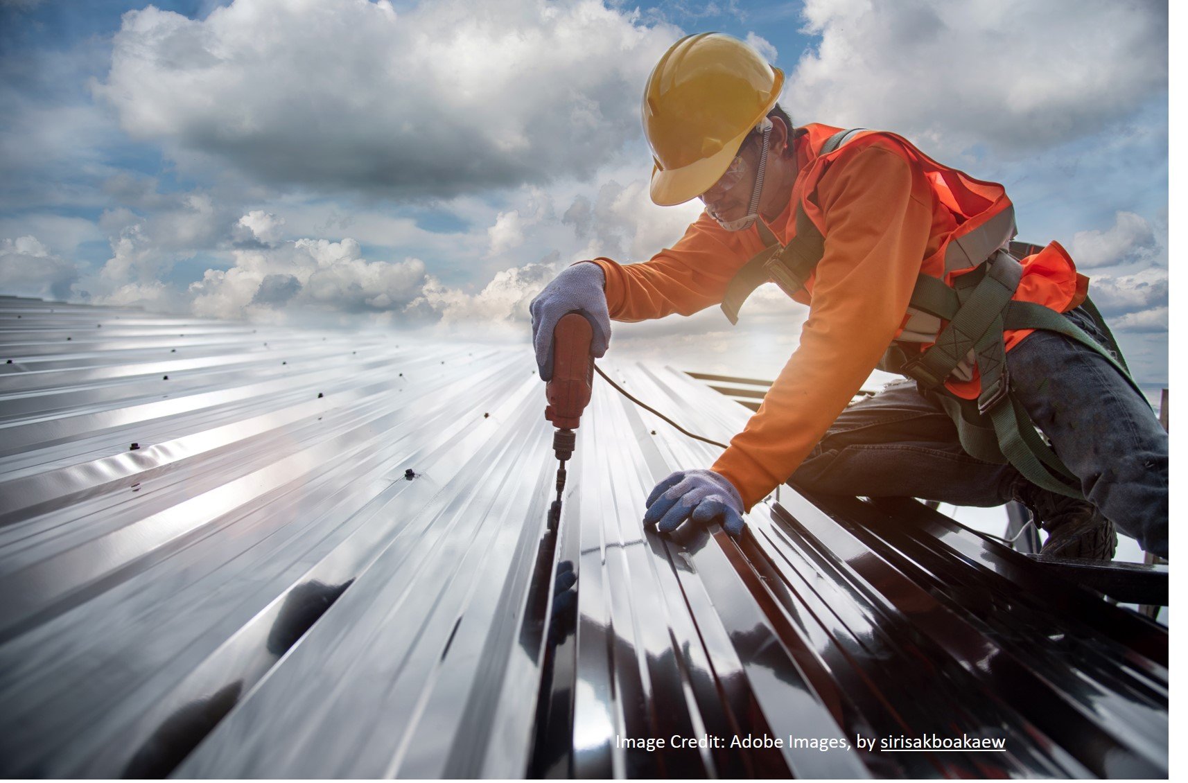 Worker in a hard hat and safety harness drilling a metal roof panel into place high above the ground.