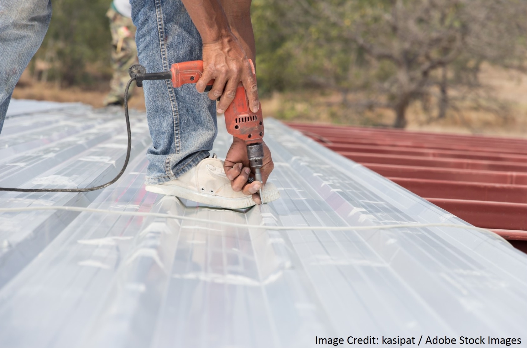 A close-up of a worker's hands drilling into a clear corrugated roofing panel beside red metal panels.
