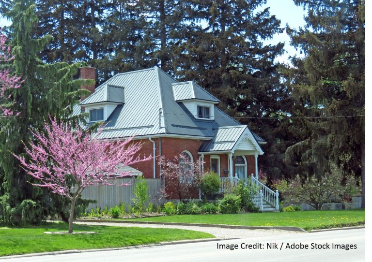 Historic red-brick home with a flared silver standing seam metal roof, dormer windows, and a decorative covered porch in spring.