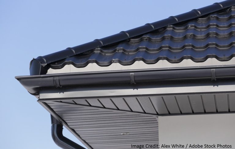 A close-up of a roof corner showing dark curved tile shingles, a metal gutter, and vented soffit panels.