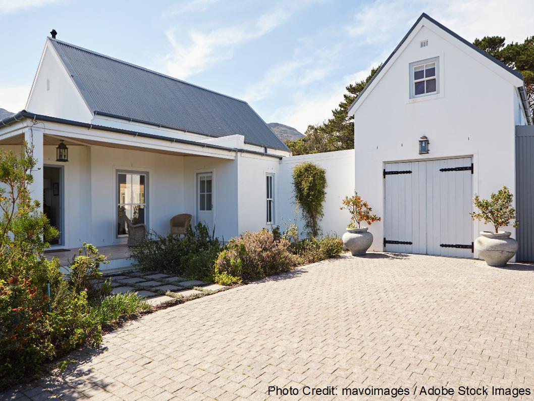 A white country-style home and attached garage feature a corrugated metal roof surrounded by lush landscaping and a cobblestone driveway.