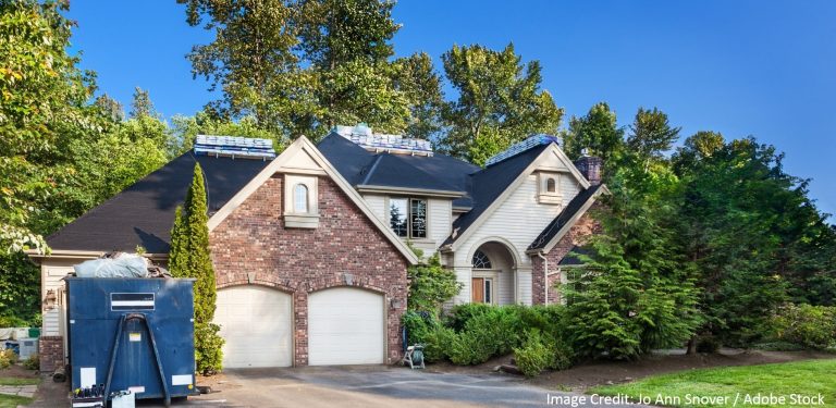 A brick and stone home with new shingle bundles on the roof and a dumpster parked in the driveway.