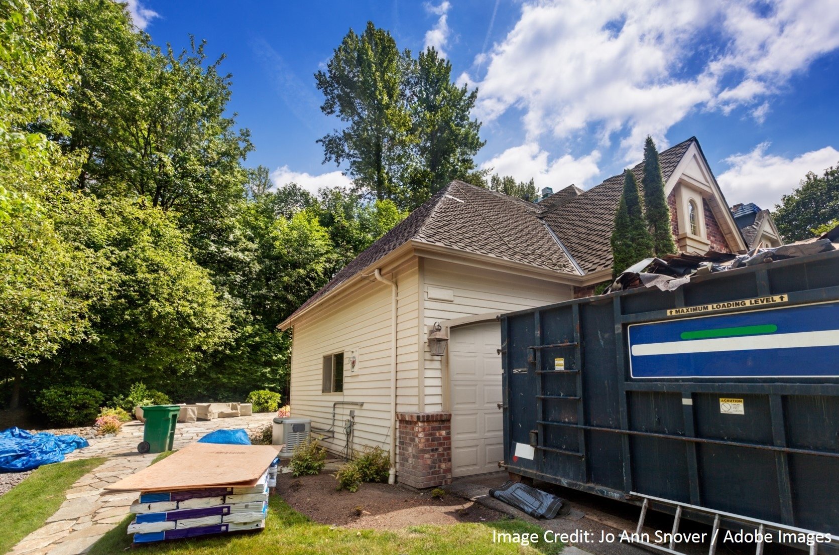 A home with a partially finished shingle roof sits beside a large dumpster and staged construction materials.