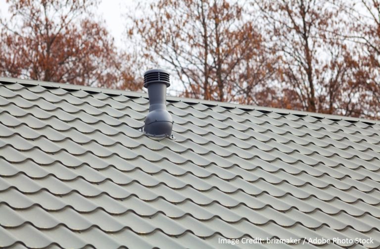 Close-up of a green tile-profile metal roof with a dark exhaust ventilation pipe against a backdrop of bare autumn trees.
