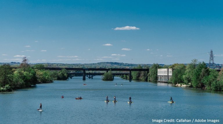 Paddleboarders and kayakers glide across a calm river near a railroad bridge beneath a bright blue sky.