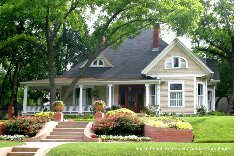 A tan Victorian-style home with a wraparound porch and dark shingle roof sits behind a colorful flower garden.