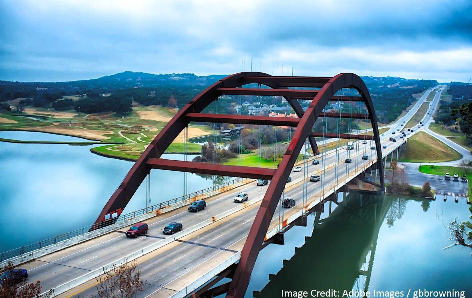 An arched steel bridge carries traffic across a calm lake surrounded by green hills and clouds.