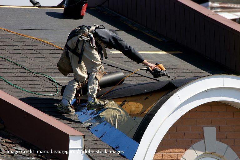 A roofer crouches on a sloped shingle roof while using a pneumatic nail gun near an arched brick dormer.