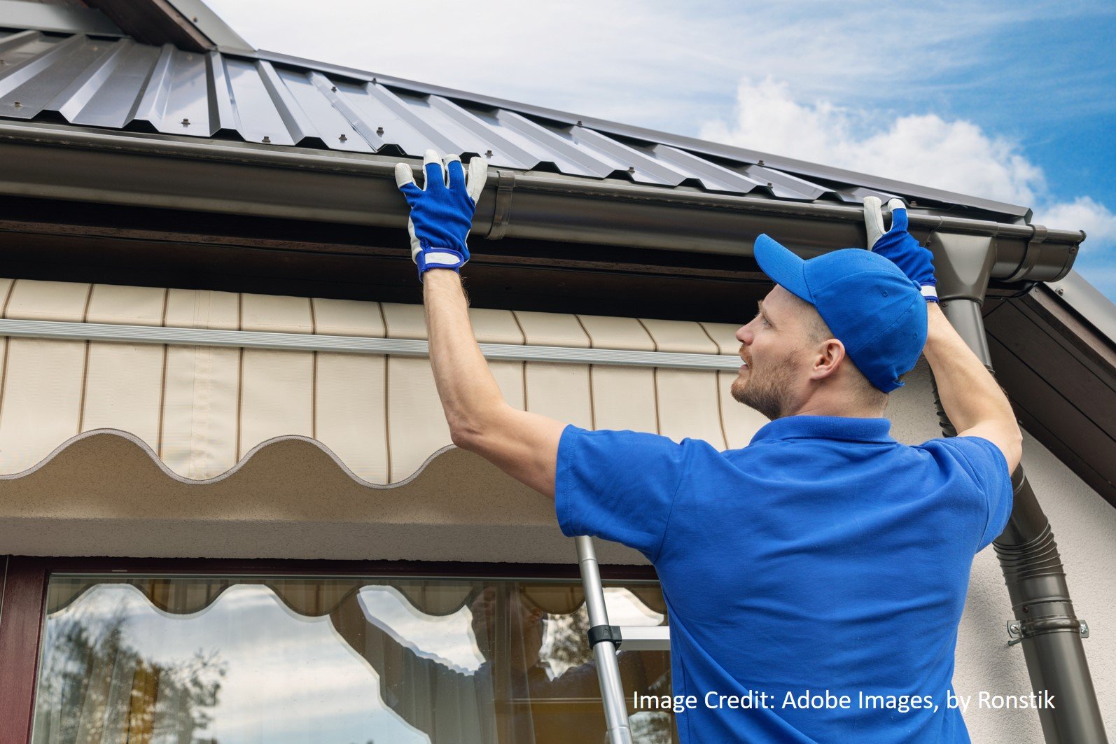 A man in a blue uniform installs gutter trim beneath a standing seam metal roof above a scalloped fabric awning.