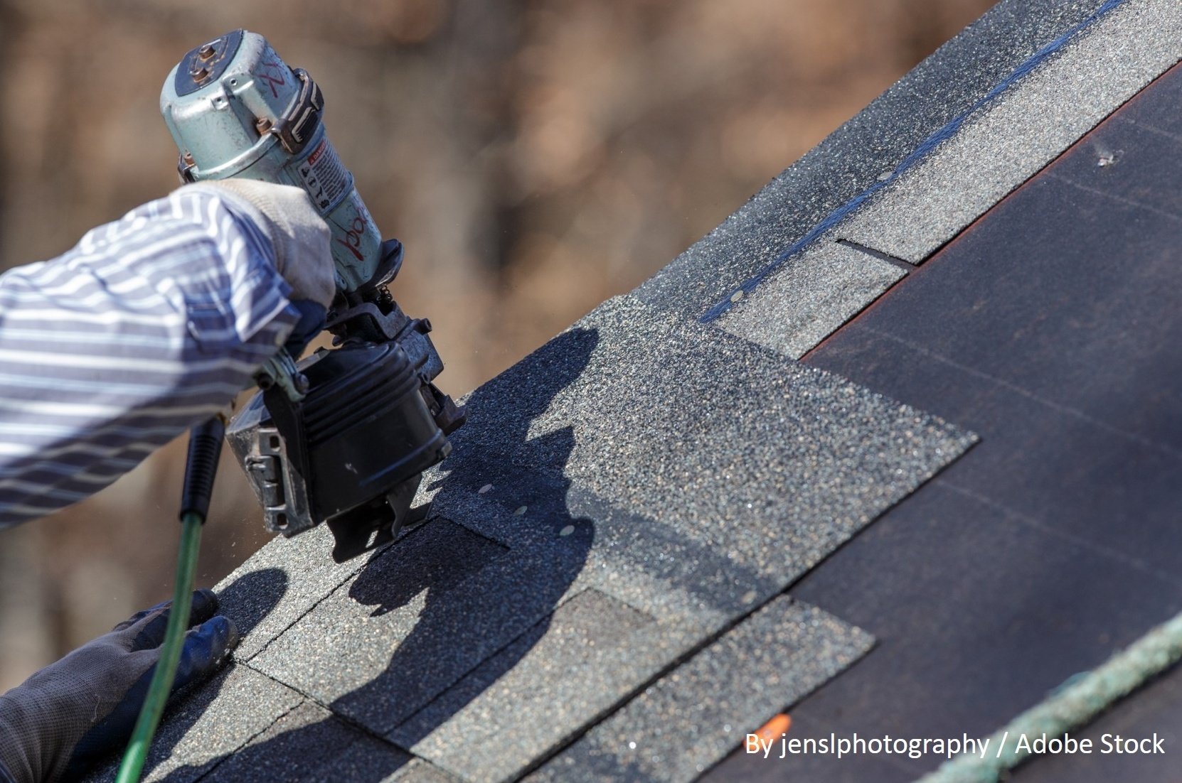 Close-up of a roofer using a pneumatic nail gun to fasten gray asphalt shingles along a sloped rooftop.