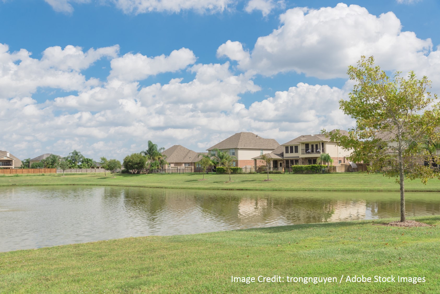Upscale homes with tile roofs sit along a calm lake shoreline beneath a bright blue sky with fluffy clouds.