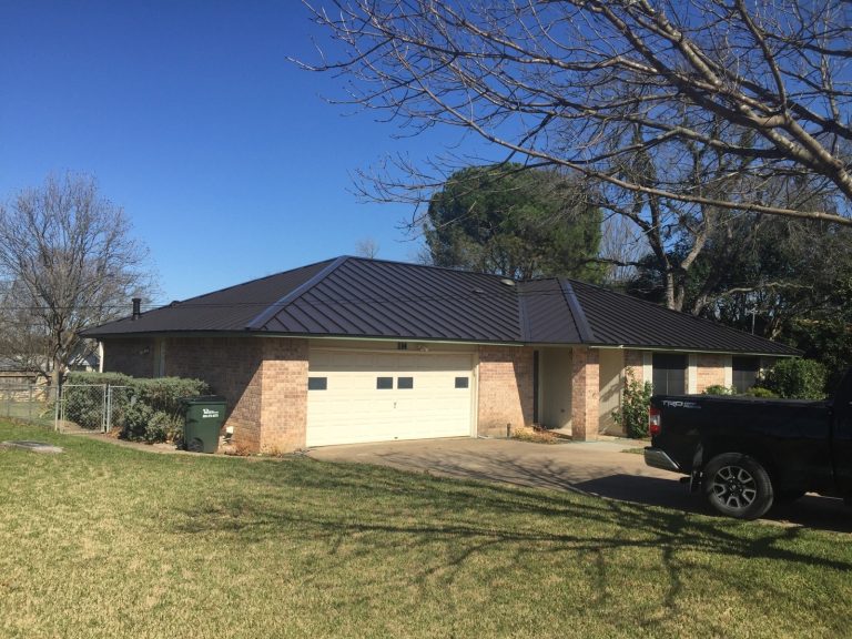 A brick ranch-style home with a dark standing seam metal roof sits behind a chain-link fence, with a truck parked in the driveway.