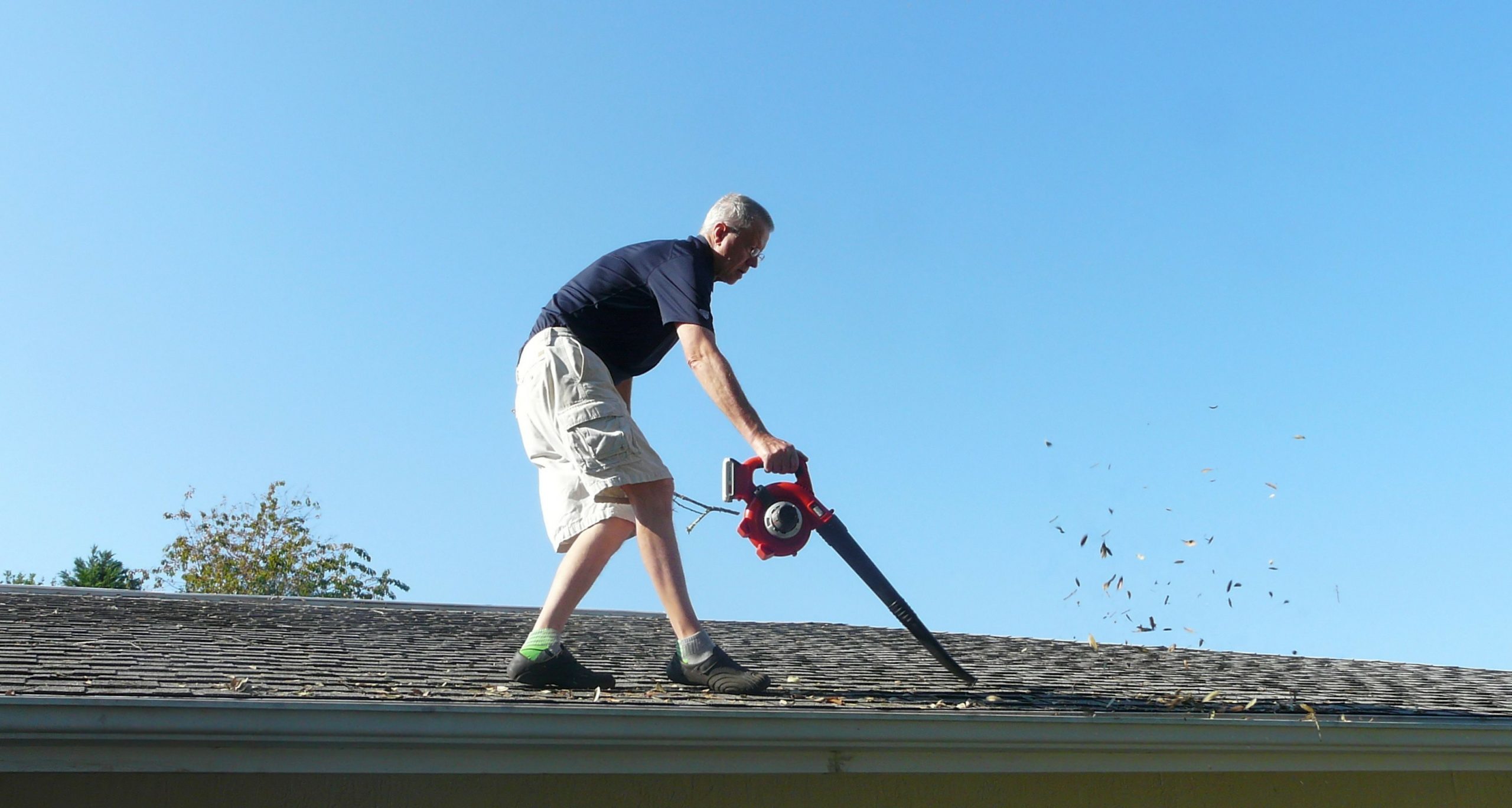 Man using a red leaf blower to clear fallen leaves and debris off a gray shingle roof under a clear blue sky.