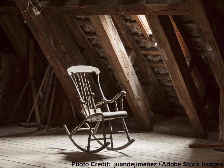 An antique wooden rocking chair sits alone in a dim attic, surrounded by exposed diagonal wooden roof rafters and beams.