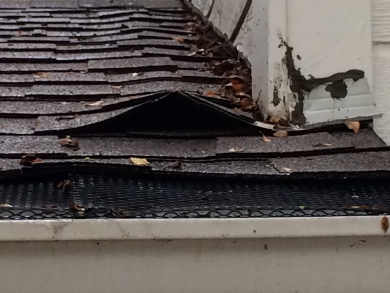 A close-up of curling, deteriorated shingles and peeling trim paint along a worn roof corner and gutter.