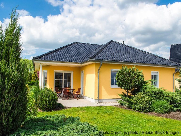 A yellow stucco home features a dark tile hip roof, surrounded by manicured shrubs and tall evergreen trees.