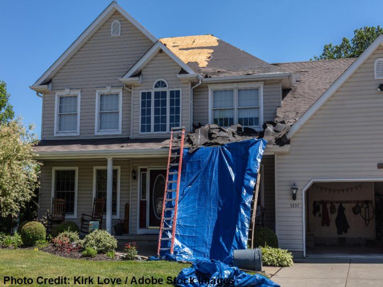 A two-story home shows an exposed section of roof decking, covered partly by a large blue tarp and a leaning ladder.