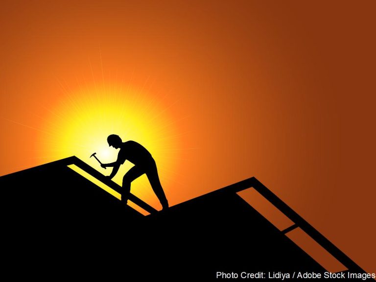 A roofer's silhouette is framed against a vivid orange sunset while hammering along the peak of a rooftop.