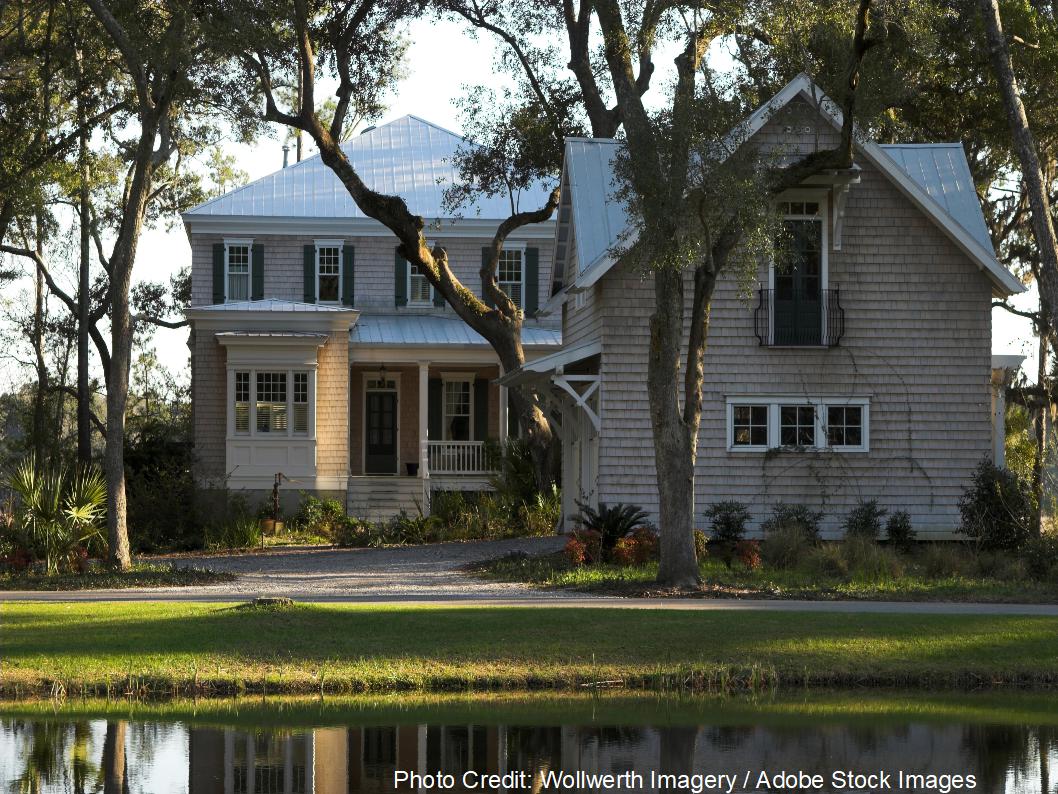 A stately two-story home with a standing seam metal roof sits beside a still pond, framed by moss-draped oak trees.