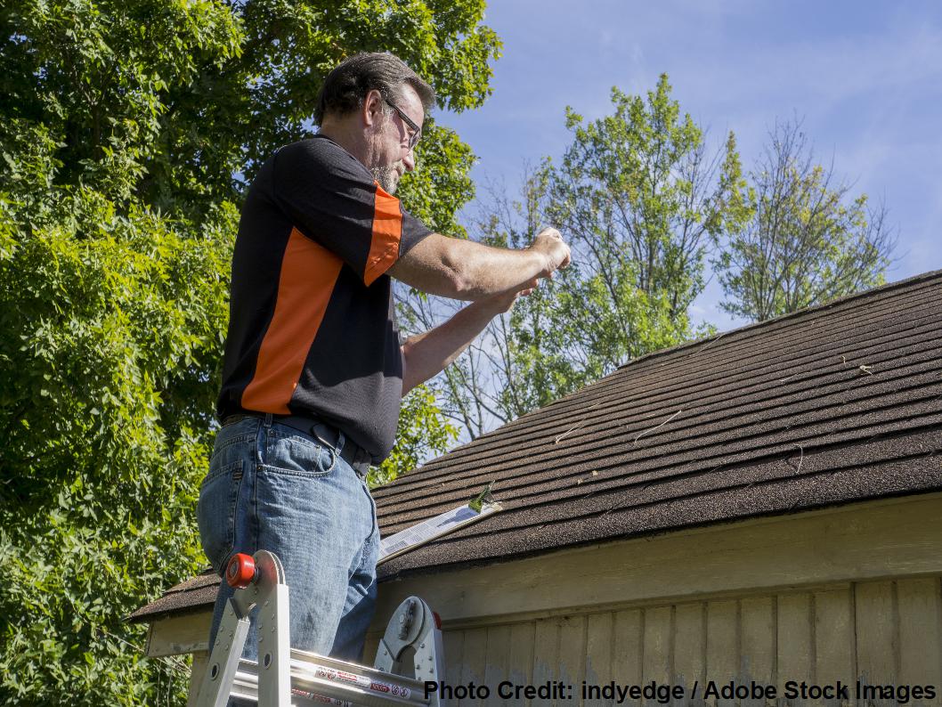 A man on a ladder examines asphalt shingles on a garage roof, surrounded by green trees under a clear blue sky.
