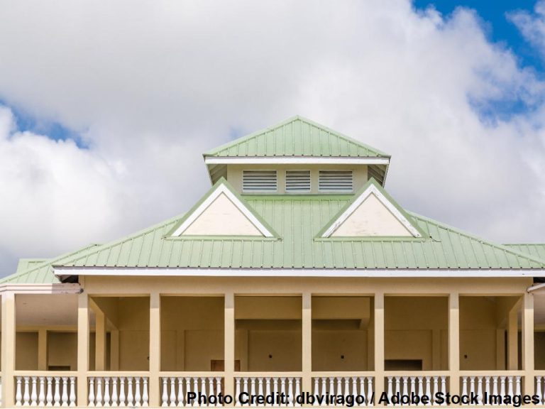 A stately colonial-style building features tall white columns and a green hip roof with dormer windows beneath a partly cloudy sky.