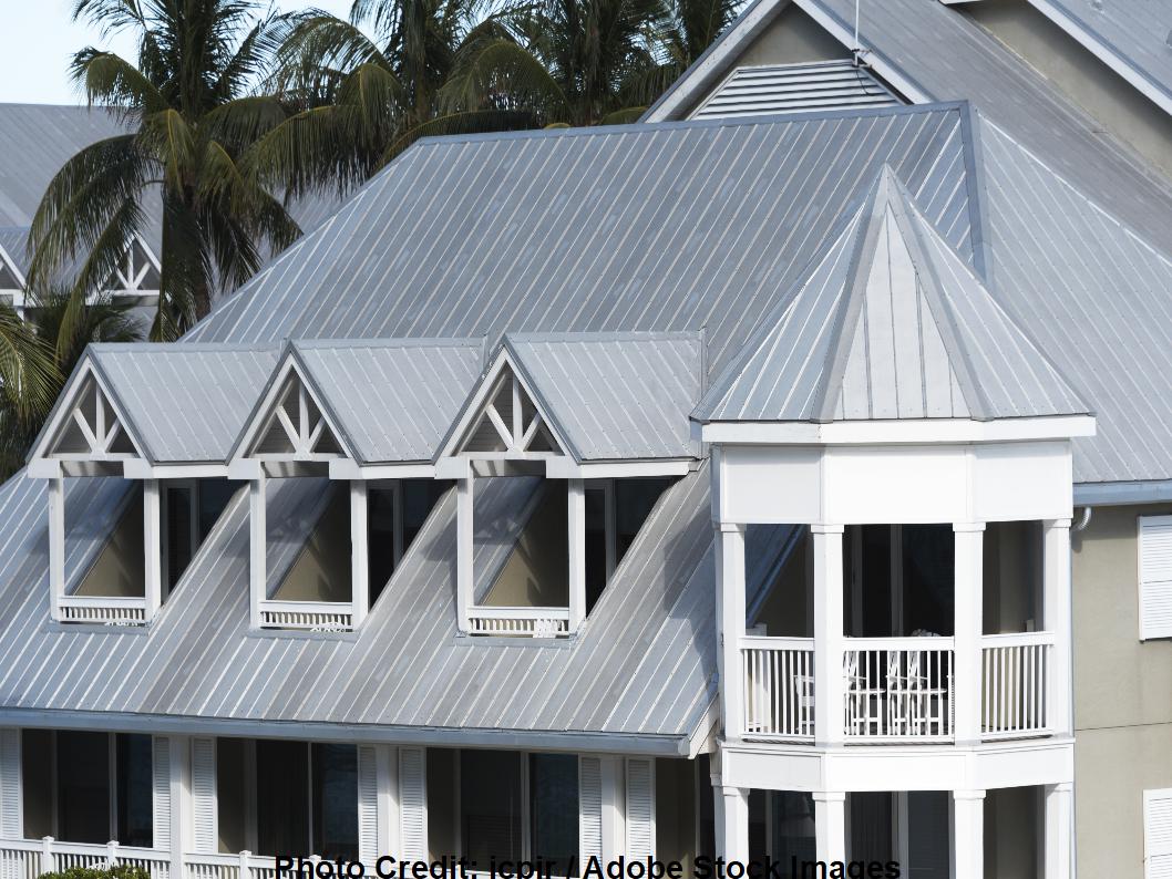 A multi-gable home features gray standing-seam metal roofing and white wraparound porches, surrounded by tall palm trees.