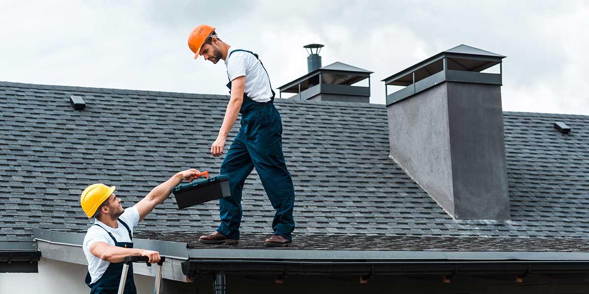 One roofer on a ladder hands a toolbox up to another standing on a gray shingle roof, both wearing hard hats.