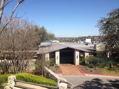 A stone building with a metal roof and arched entrance overlooks a wooded hillside beneath a clear blue sky.