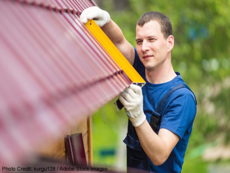 A roofer wearing gloves carefully measures a red metal roof panel using a yellow tape measure during routine maintenance.