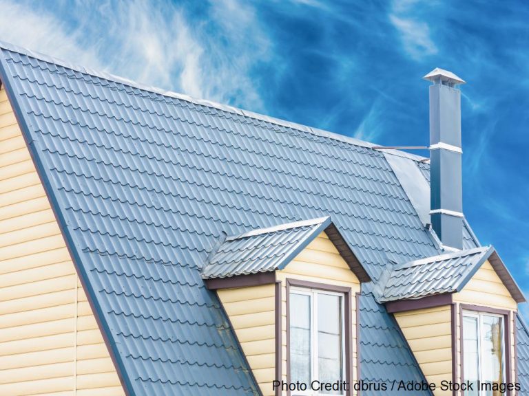A blue-gray tile-pattern metal roof rises above tan siding, with two dormer windows and a tall metal chimney vent.