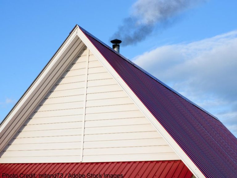 A dark red metal roof gable with white siding features a vent pipe releasing smoke into a blue sky.
