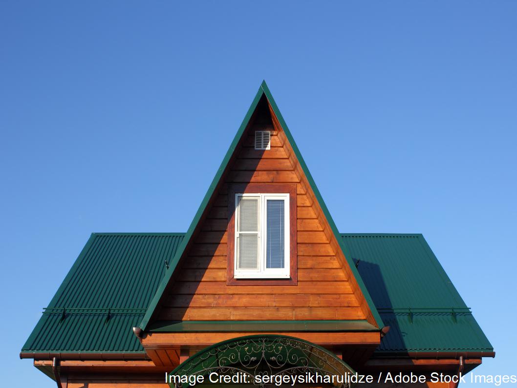 A wooden A-frame gable with a white window rises beneath a green corrugated metal roof and clear blue sky.