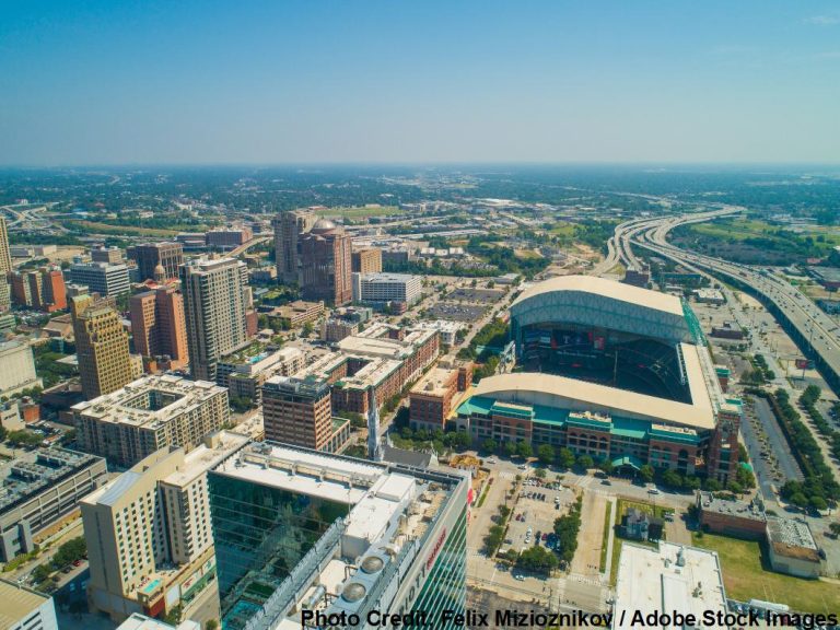 An aerial view captures a downtown skyline with a retractable-roof baseball stadium and a highway curving through the city.