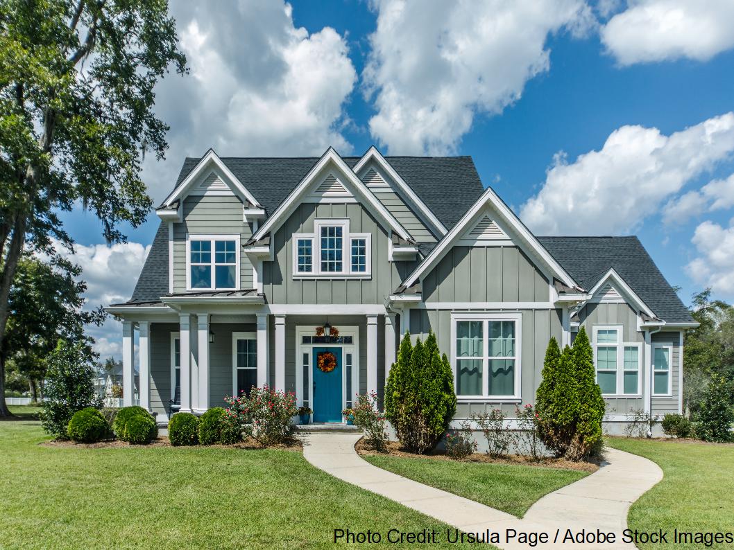 A two-story gray craftsman-style home features multiple gables, white trim, and a dark asphalt shingle roof above a landscaped yard.