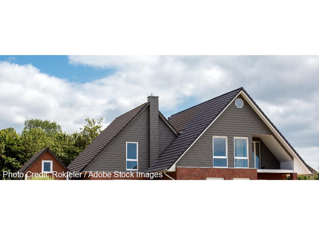 A home features a steep dark tiled gable roof, a round accent window, and a brick lower exterior beneath a cloudy sky.