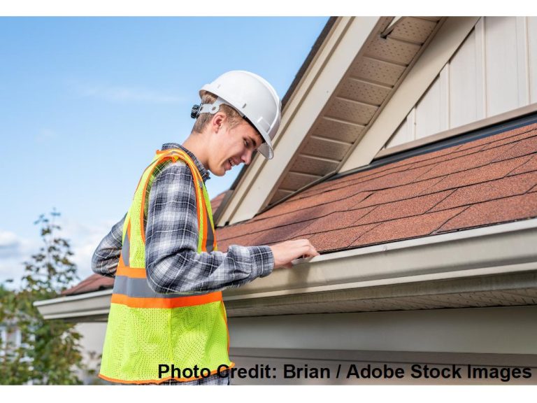 A roof inspector wearing a hard hat and safety vest closely examines the shingle edge and gutter along a home's roofline.