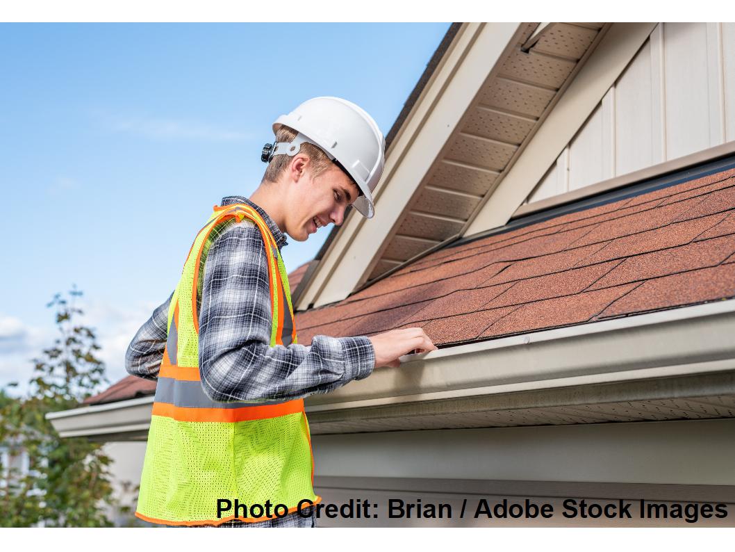 A roof inspector wearing a hard hat and safety vest closely examines the shingle edge and gutter along a home's roofline.